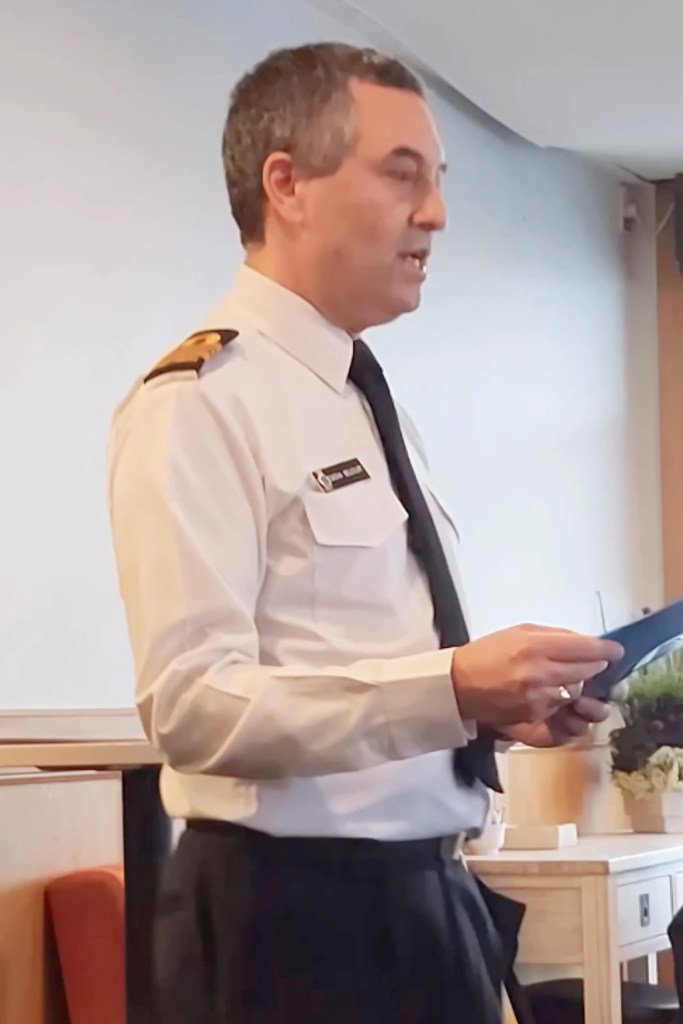 In the image, there is a man standing indoors, presenting or reading from a document. He appears focused and professional, dressed in a light-coloured shirt with dark trousers. On his left shoulder, he wears a military-style epaulette with a single thick and a single thin gold stripe, indicating a rank that could be associated with naval or maritime services. A badge is visible on his left breast pocket, though the details are not clear. The background is neutral and unfocused, allowing the subject to stand out. His attire and demeanor suggest a formal or ceremonial context, perhaps related to a naval or maritime organization's event.