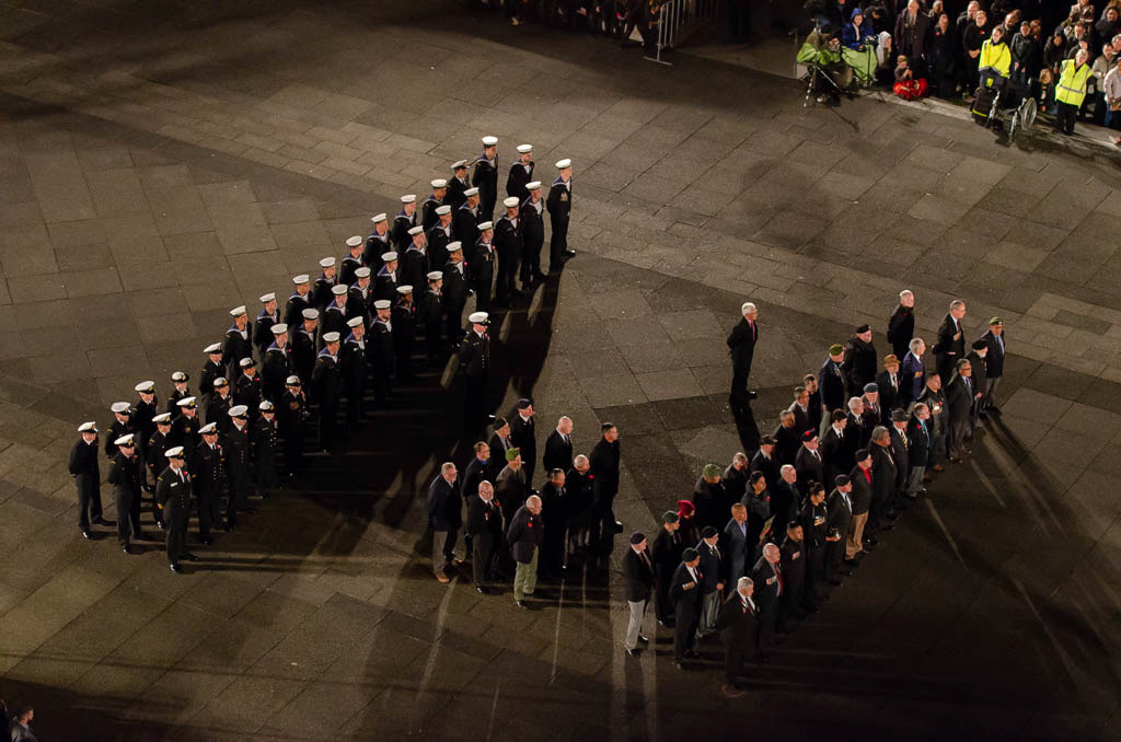 The image captures a ceremonial gathering at dawn, illuminated by ambient light. A formation of uniformed navy personnel, is arranged in a block pattern on a paved area. They are wearing white peaked caps, and their dark uniforms contrast with the light-colored ground. To the right, a group of veterans, some wearing medals and traditional red poppy pins, stands in rows. Observers are visible in the background, behind barriers, indicating this is a public event. The occasion appears to be somber and respectful, characteristic of a commemorative service such as Anzac Day, where military and civilians come together to remember and honour those who have served.