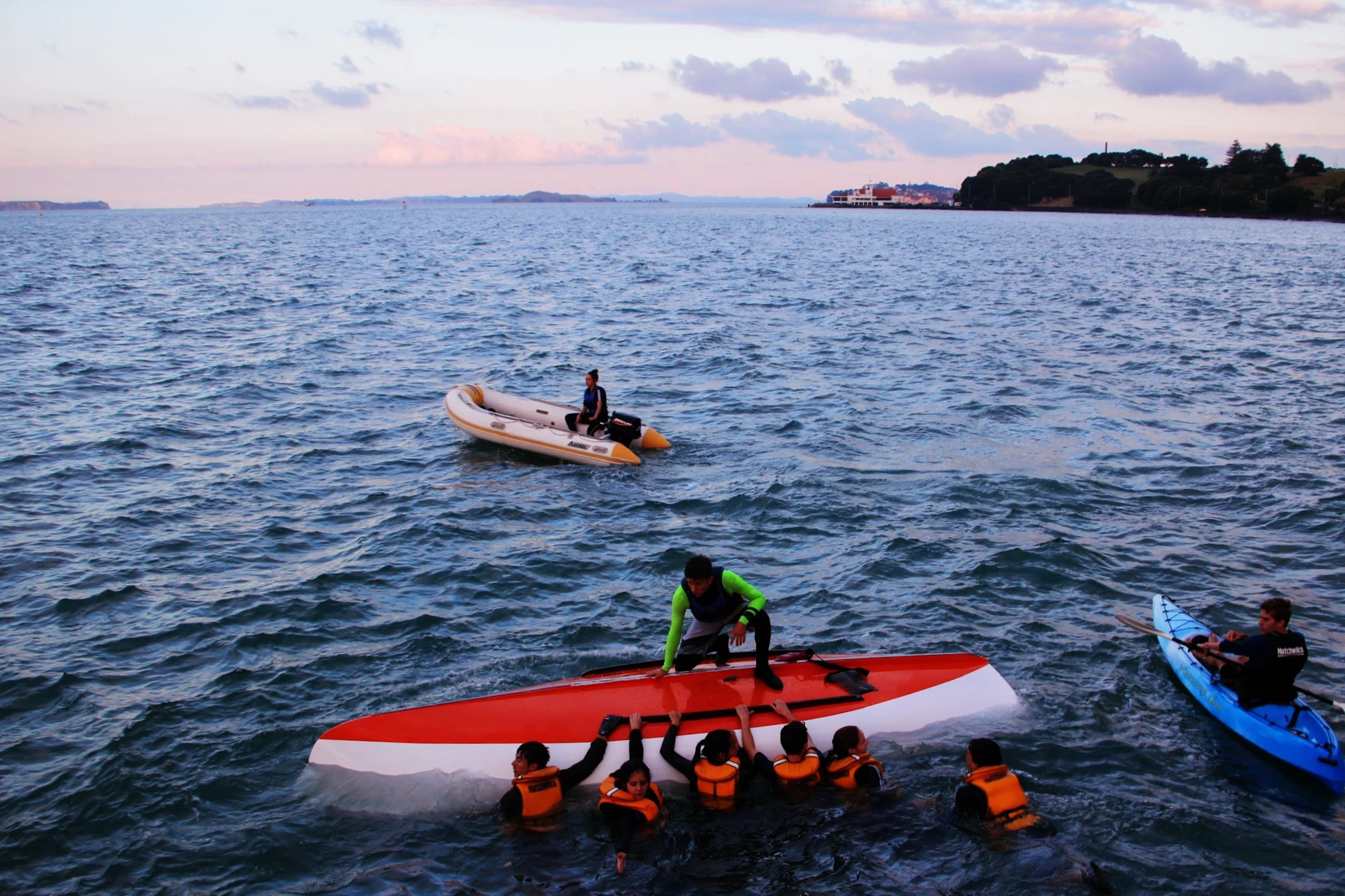 In the image, we see an outdoor water training exercise in progress during twilight. A group of individuals in orange life jackets are in the water around a capsized sailboat, with one person on the hull, actively participating in a capsize recovery drill. To the left, a person on an inflatable dinghy oversees the exercise, while another individual on a kayak is nearby, ready to assist. The water is a deep blue with gentle waves, and the sky above is a mix of soft pink and blue hues, indicative of either dawn or dusk. In the background, there's a hint of land with trees and structures, suggesting this activity is taking place close to shore. The scene is lively with the effort of the training and the calmness of the surrounding nature.