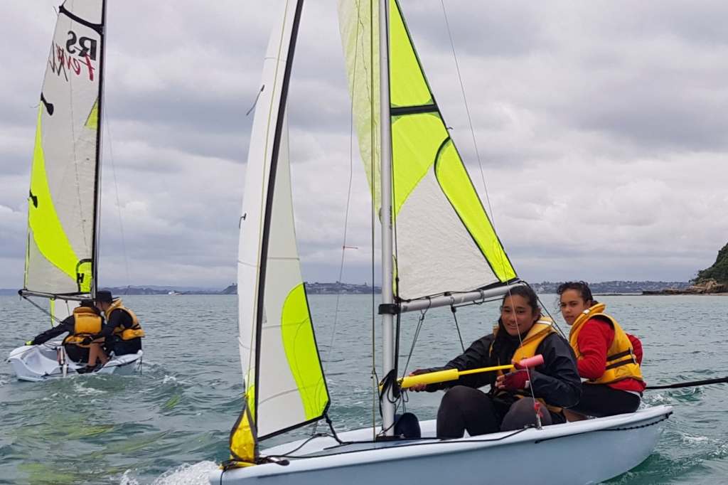 The photograph features two sailboats gliding over the water, with the foremost vessel captained by two sailors. The sailors are wearing bright yellow life jackets for safety and are attentively navigating their craft. The main focus is on the crew member in the stern, who holds the tiller extension and seems to be steering, while her companion looks on from the bow. Both have a backdrop of the coastline and a calm sea that stretches to the horizon. The sails of both boats are a striking combination of white and fluorescent green, catching the eye and standing out against the overcast sky. The expression on the faces of the sailors is one of concentration and confidence, embodying the essence of seamanship.