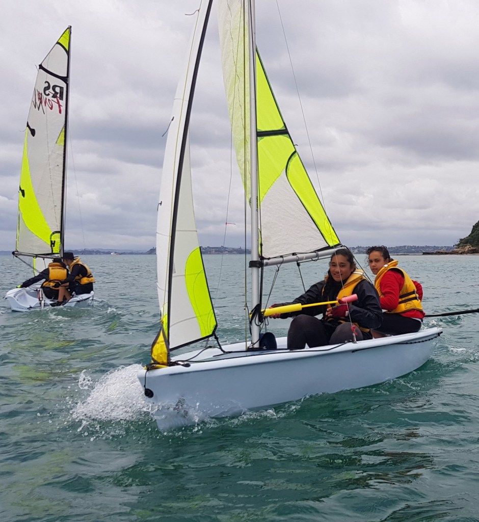 The image captures a vibrant sailing scene with two small sailboats. Each boat is manned by two individuals wearing life jackets, one in yellow, the other in red. They are actively engaged in sailing, with the closest pair looking towards the camera, navigating a boat with a white hull and sails adorned with bright yellow and white patterns. Water sprays up at the bow of their boat, illustrating their swift movement through the water. The background features a calm sea and a coastline dotted with trees and buildings. Overhead, the sky is a mix of blue and grey, suggesting a brisk but pleasant sailing condition.