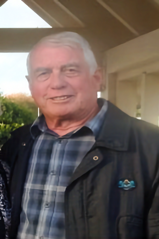 The image shows an older gentleman with a cheerful expression. He is wearing a casual, layered outfit consisting of a dark jacket over a plaid shirt in shades of grey and blue. The jacket features a distinctive badge on the left chest. His silver hair is complemented by the warm, outdoor lighting that suggests the photo was taken around dusk. The background is softly blurred, focusing attention on his face, and it appears to be an outdoor residential setting.