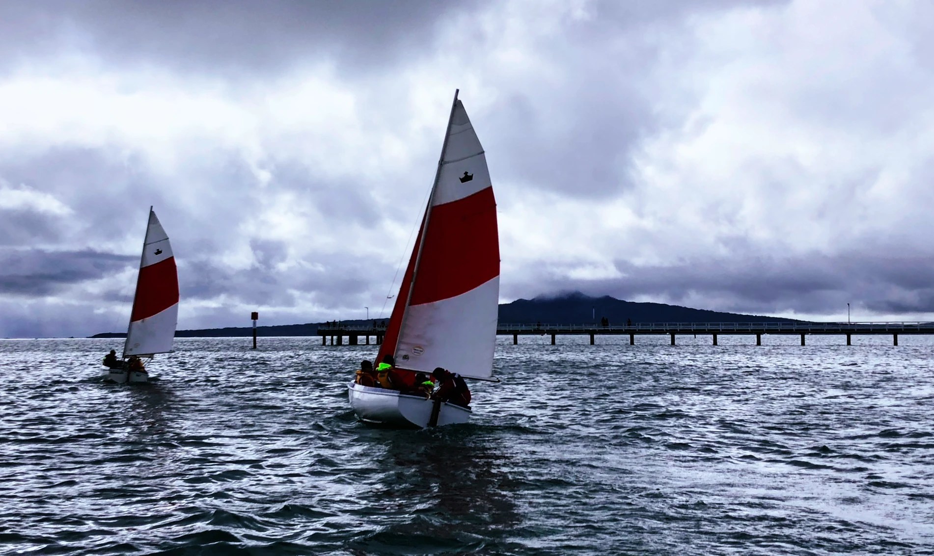 This image features two small sailboats with white and red sails navigating choppy grey-blue waters. The sailors, clad in bright life jackets, are seated closely as they manage their vessels. Overcast skies loom above, filled with heavy clouds that promise inclement weather, yet the scene retains a dynamic sense of adventure and activity. In the background, a long pier extends into the water, leading to a hazy landmass that fades into the distance, creating a sense of depth and scale. The overall impression is of a brisk sailing day where sailors are undeterred by the moody weather, embracing the challenge with enthusiasm.