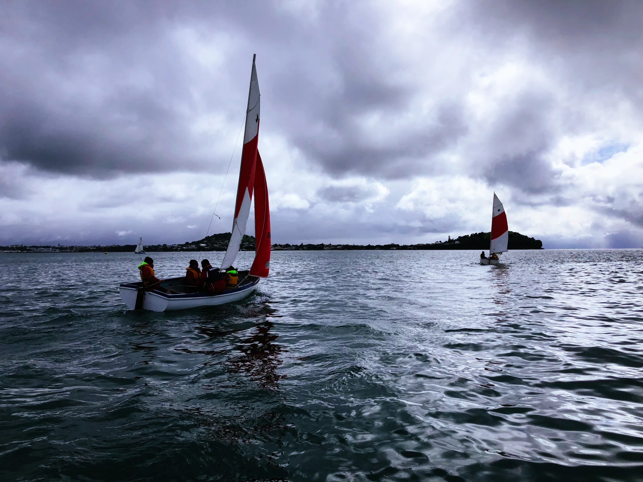 The image features two sailboats on an expanse of water, under a dynamic sky filled with heavy, textured clouds. The closest sailboat, with its white sail marked by a red chevron, is crewed by sailors in vibrant life jackets. They seem to be in a moment of calm, with the boat gently tilting with the wind. The water reflects the grey tones of the sky, suggesting a somber or overcast day. In the distance, a headland can be seen, adding a sense of scale and location to the scene. The photograph conveys a quiet anticipation, with the sailors poised for action, yet the atmosphere is one of tranquillity.