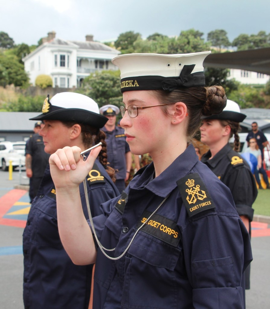 The photograph displays a group of uniformed cadets at an outdoor ceremony. In the foreground, a cadet is captured mid-gesture, her arm raised and hand positioning a boatswain's call near her lips, which suggests she is about to issue a command. She wears a navy blue uniform with the insignia of the Sea Cadet Corps, and her head is adorned with a traditional white sailor's cap marked with the word 'WAIREKA.' Her focused gaze and poised demeanor reflect discipline and readiness. In the background, other cadets in similar attire stand at attention, with a hint of a crowd and greenery behind them, indicating a formal event.