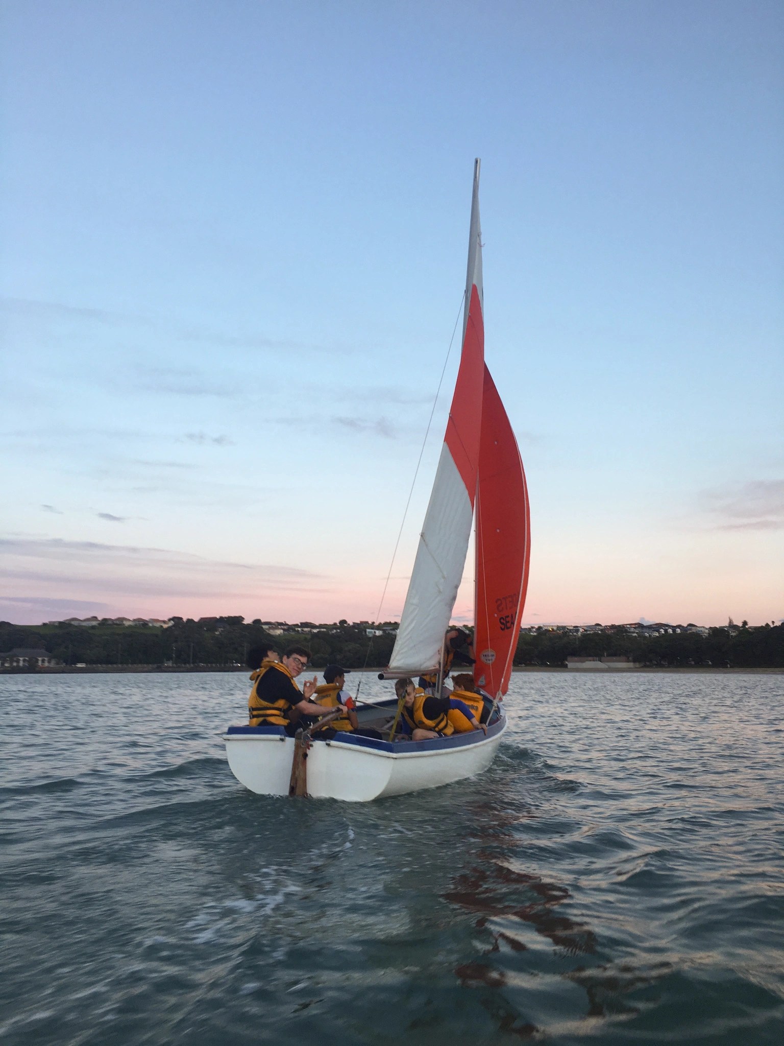 This image depicts a sailboat at dusk, its white and red sail billowing in the wind as it glides through the water. The boat is crewed by individuals wearing bright yellow life jackets, indicating a focus on safety. The sailors appear engaged and are working together to navigate the vessel. The sky above is painted with the soft pastel hues of the evening, transitioning from the warmth of the day to the cooler tones of night. The water reflects the fading light, creating a serene and peaceful maritime scene. In the background, the silhouette of a coastline can be discerned, suggesting the boat is not far from shore. The overall atmosphere is one of calm and enjoyment of sailing during the quiet of twilight.