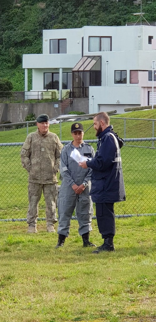 The image shows three individuals in a discussion in what appears to be a field training environment. To the left stands a person in military camouflage attire and a beret, in the centre is someone in a grey jumpsuit with a cap, denoting a naval cadet, and to the right is a person in a dark blue jacket with reflective elements, indicative of a naval instructor. They are engaged in conversation, possibly about the training exercise at hand. In the background, a modern, multi-story residential building can be seen, providing a contrast between the calm domestic setting and the focused, professional exchange taking place in the foreground. The lush green grass and overcast sky suggest an outdoor training scenario during cooler weather.