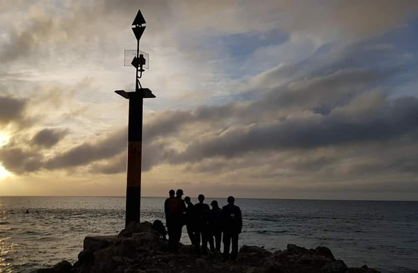 This image captures a silhouette of a group of individuals gathered near a navigational marker at dusk. The marker is a tall pillar with a triangular top, standing prominently against the twilight sky. The sky is painted with hues of orange and blue, with the sun just below the horizon, casting a warm glow that softly illuminates the clouds. The sea is calm, stretching into the horizon and reflecting the fading light. The figures are standing on rocky terrain, suggesting a coastal location. The overall mood of the image is serene and contemplative, highlighting the beauty of the sea at sunset.