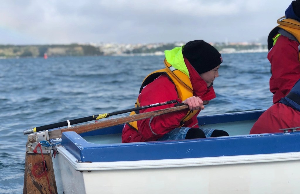 The image captures a close-up moment of a young sailor at the helm of a small boat. The individual is clad in a red life jacket with a high-visibility yellow hood, a black beanie, and is actively engaged in steering, gripping the tiller with focus. The backdrop is a blurred seascape with hints of a distant shoreline, suggesting the photo was taken in a dynamic marine setting. The concentration on the sailor's face and their firm grip on the tiller reflect the attentiveness required to navigate the waters, while the attire indicates cool or potentially adverse weather conditions. The angle of the shot, with the sailor's face partially turned, adds a sense of immediacy and immersion into the sailing experience.