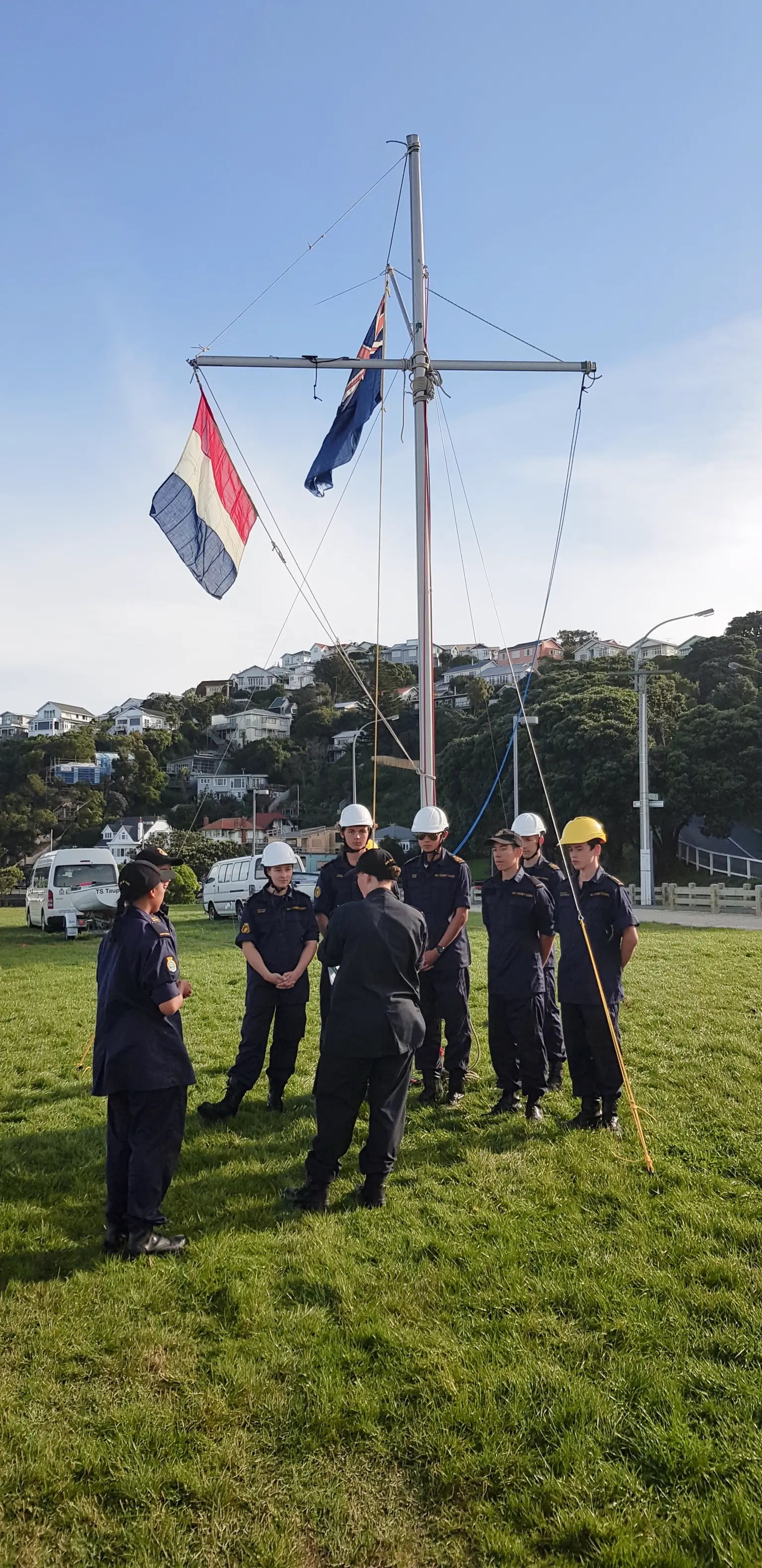 The image captures a group of cadets engaged in maritime training, centered around a mast construction. The cadets are attired in various uniforms, some in dark blue with white caps and others in safety helmets. They seem to be in the midst of a hands-on learning session, with one cadet handling a wooden pole and others standing in a semi-circle receiving instruction. The background features a clear sky and the serene residential backdrop of a hillside community, suggesting the location might be a coastal area. Flags, including what appears to be the New Zealand Ensign, fly from the rigging. The scene is one of teamwork and focus, with the cadets' attention directed towards their instructor, demonstrating the learning and discipline that are integral to cadet training.