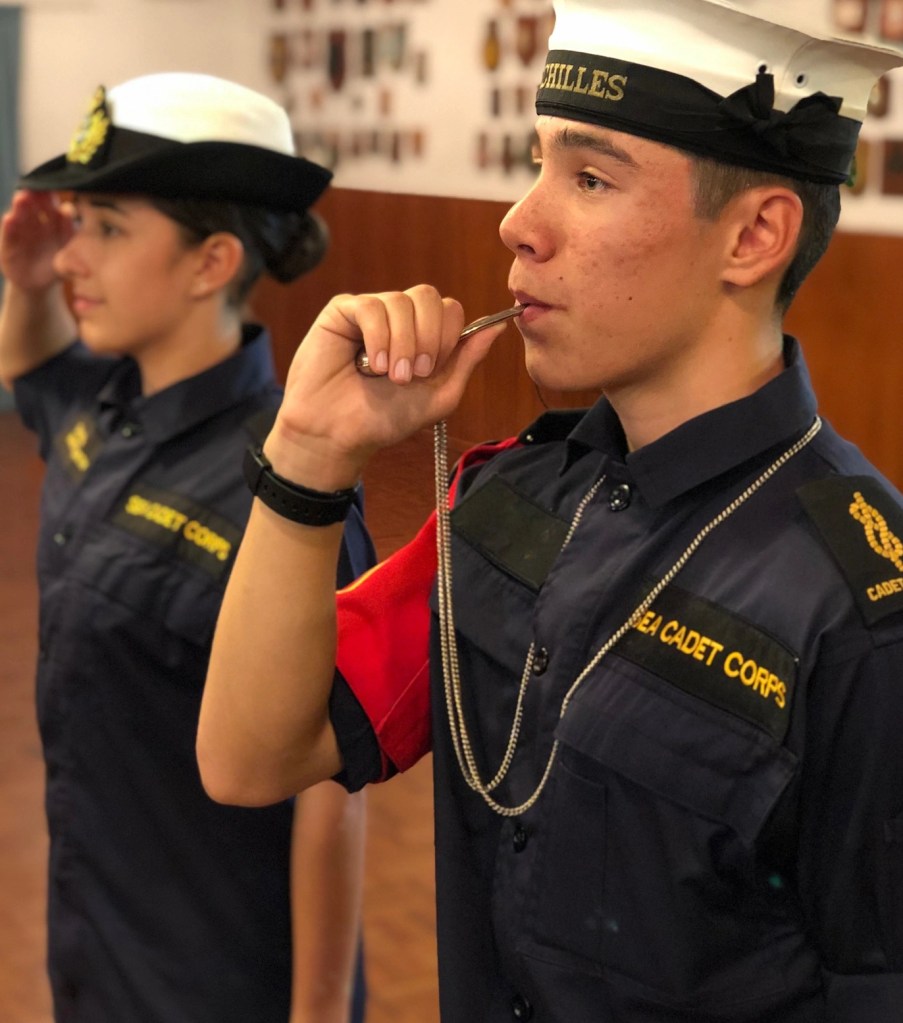 The image depicts two cadets in naval uniforms participating in a ceremonial activity indoors. The foreground shows a male cadet in profile, wearing a dark blue shirt with the words "Sea Cadet Corps" and a cap emblazoned with "Achilles." He is blowing a boatswain's call (a whistle used in naval communications) held by a chain in his left hand, a traditional naval practice. Behind him, partially out of focus, is a female cadet saluting, also in a dark blue uniform with a white peaked cap. The backdrop includes a wall displaying plaques or badges, suggesting the setting may be a naval or maritime institution. This scene conveys a sense of tradition, discipline, and the continuation of naval customs within the cadet training environment.