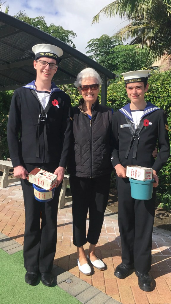 The image features two cadets and an older woman standing outside on a sunny day. The cadets are dressed in naval cadet uniforms, with white hats labeled "T.S. Achilles" and dark jackets with white belts. Both have red poppies pinned on their jackets, indicating the photo was likely taken around Anzac Day or Remembrance Day. The cadet on the left holds a collection box labeled "RSA," which stands for the Returned and Services' Association, an organization supporting military veterans in New Zealand. The cadet on the right holds a donation bucket. The woman in the center, likely a supporter or perhaps a veteran herself, wears a black puffer vest, black trousers, and sunglasses, with a poppy on her vest. They are all smiling, suggesting a positive atmosphere during a commemorative event. The setting includes a covered walkway and a backdrop of trees and a residential area. The image captures a moment of intergenerational connection and the continuation of traditions honoring military service.