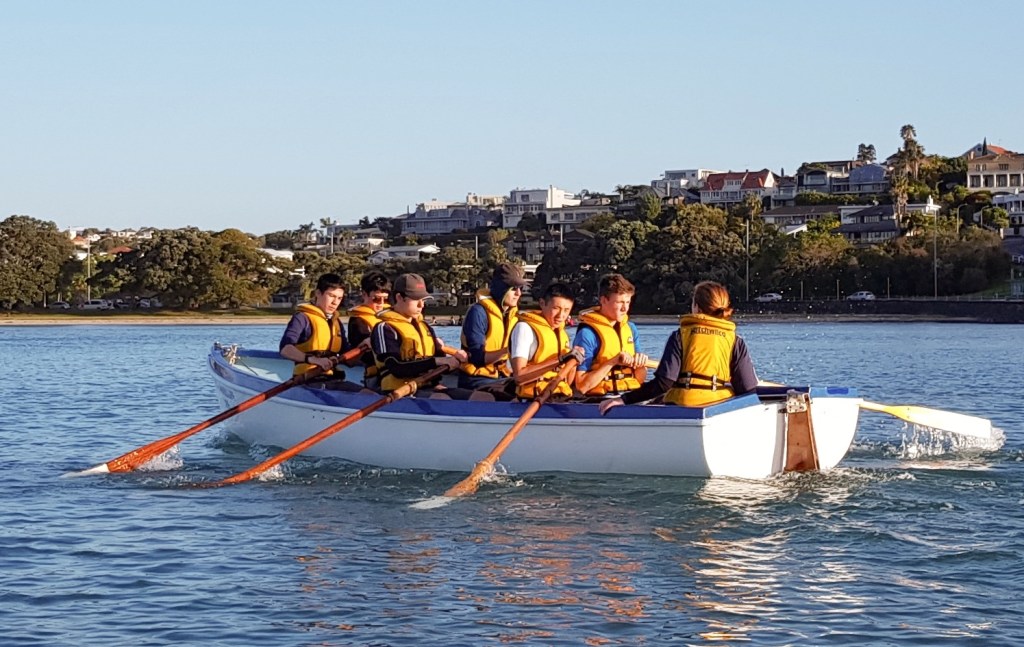 The image features a team of rowers in a large, white boat on a tranquil expanse of water, bathed in the warm glow of the setting sun. Each rower is adorned with a vivid orange life jacket, and they are caught mid-stroke, their oars dipped in the water, creating ripples. The rowers are concentrated and synchronised in their effort. In the background, the shoreline is adorned with a variety of trees and a sprinkling of residential buildings, with the clear blue sky above them. The peaceful evening light lends the scene a serene and amiable atmosphere.