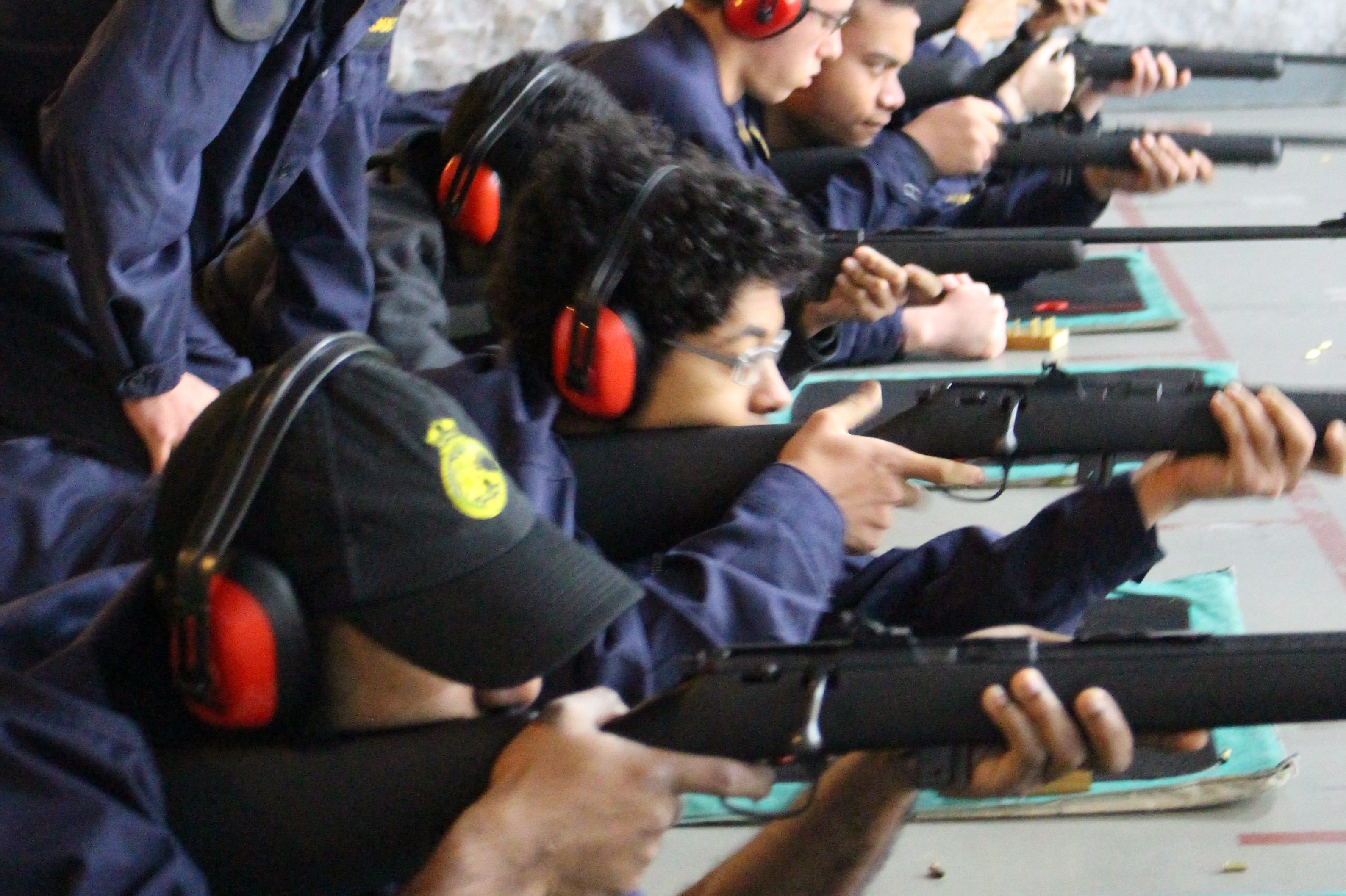 The photograph captures a line of individuals engaged in a precision shooting exercise at an indoor range. They are dressed in dark blue uniforms, each with a pair of red ear defenders for protection against the noise. The participants are lying prone on green mats, each aiming a black, bolt-action rifle down the range. Their postures suggest focus and discipline as they take aim. The foreground shows a small wooden block holding a set of rounds, indicating the live nature of the exercise. The environment is functional, with a concrete wall in the background, which emphasises the controlled and safe setting of the activity.