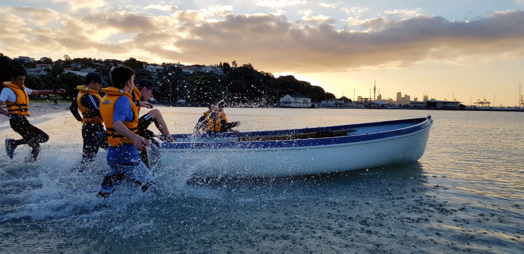 This dynamic image captures a group of individuals participating in what appears to be a team-building activity, possibly at dawn or dusk. They are clad in orange and blue life jackets and are in various stages of exiting a large blue and white boat onto the shore. Splashes of water rise around them as they make their way through the shallow water, with the city skyline silhouetted against the glowing sky in the background. The setting sun casts a warm golden light over the scene, contrasting with the urban silhouette that includes a distinctive tower, hinting at a coastal city's waterfront.