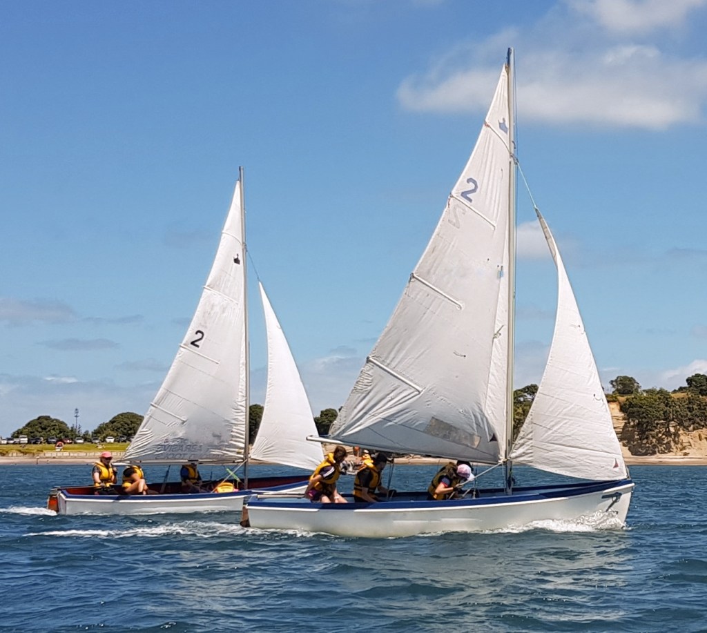 A vibrant photograph captures two sailboats in action on a sunlit day. Both vessels boast crisp white sails, with the number '2' prominently displayed, indicating their participation in a regatta. The boats are manned by teams dressed in yellow life jackets; some team members are seated while others are actively working the sails. The boats are tilting slightly due to the wind and momentum, cutting through the glistening blue waters. In the background, a serene shoreline with verdant trees under a clear blue sky completes the picturesque scene.