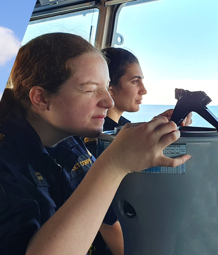 The photo shows two individuals inside a ship's bridge, concentrating on navigation tasks. The person in the foreground, with eyes closed, seems to be visualising or memorising, while the person in the background holds a pair of dividers over a chart, indicative of plotting a course. They are both wearing dark blue uniforms with 'CADET CORPS' embroidered in gold, signifying their roles and affiliation. Natural light from outside the bridge illuminates the scene, suggesting a bright day at sea. The focus and dedication on their faces reflect the seriousness with which they approach their duties in maritime navigation.