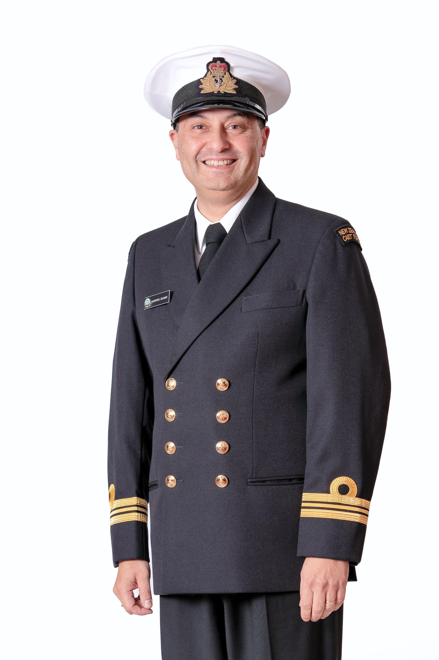 The image is a formal portrait of a man dressed in a naval uniform. He stands tall and smiles at the camera, exuding a sense of pride. The uniform is dark, with a double row of gold buttons and three gold stripes on the cuffs, signifying rank. On his left breast is a name tag, and on his right, a badge with intricate embroidery, indicative of his service and position. His head is adorned with a white peaked cap with a cap badge. The background is plain and white, putting the entire focus on his attire, stature, and pleasant demeanor.