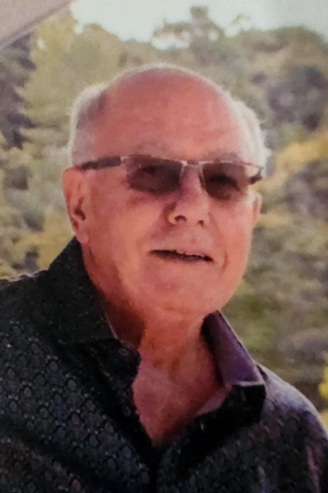 The image is a close-up portrait of an elderly gentleman with a genial smile. He is wearing sunglasses and a dark patterned shirt with a collar, suitable for a warm, sunny day. The gentleman has a clean-shaven face, and his hair is neatly cropped. In the background, there is a hint of a natural setting that appears to be lush and green, likely indicating an outdoor environment. The man's cheerful expression and relaxed demeanor suggest he is enjoying a pleasant day outside.