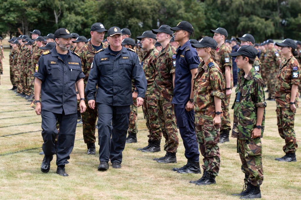 The image shows a group of military personnel in various uniforms assembled outdoors. In the foreground, there are individuals in dark naval service uniforms with distinctive cap badges, standing out against a larger formation dressed in camouflage combat uniforms. The formation appears to be arranged in neat rows, indicative of a military parade or inspection. The environment suggests an open field or parade ground, with trees and overcast skies in the background. The focus and discipline of the group are evident as they stand at attention. This scene typically represents an organized military event, possibly involving different branches of the armed forces, and reflects the formality and structure of such occasions.