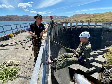 This photo depicts a sunny day with a clear blue sky and a body of water in the background, possibly a reservoir or dam. Two individuals in protective gear are engaged in an abseiling activity. The person on the left stands on a concrete ledge, wearing a dark navy uniform with cap, and is handling the rope and belaying devices, ensuring the safety of the abseiler. The second person, dressed in olive green military fatigues and a white helmet, is in a seated position against a steep concrete dam wall, secured with ropes, ready to descend. A coiled green rope lies on the ground, indicating preparations for a controlled descent. The scene is indicative of a training exercise, focusing on skill development in rappelling and the use of safety equipment in a high-angle environment. The open setting and specialized equipment suggest a rigorous and professional approach to the activity, emphasizing safety and technique in an outdoor adventure or rescue training scenario.