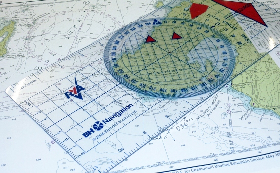 The image displays a navigational chart used for maritime purposes. The chart contains various topographical details and depth measurements critical for safe sea navigation. Placed atop the chart is a circular protractor, also known as a course plotter, which is transparent with a blue grid and measurements in degrees along its circumference. It is used for charting courses and bearings in navigation. Two red triangles, presumably course markers, are placed on the protractor, indicating specific angles or directions. The chart and the protractor together are tools used in boating education to teach the principles of maritime navigation. The logo on the plotter reads "BH Navigation". This equipment is essential for those learning to navigate water vessels accurately and safely.
