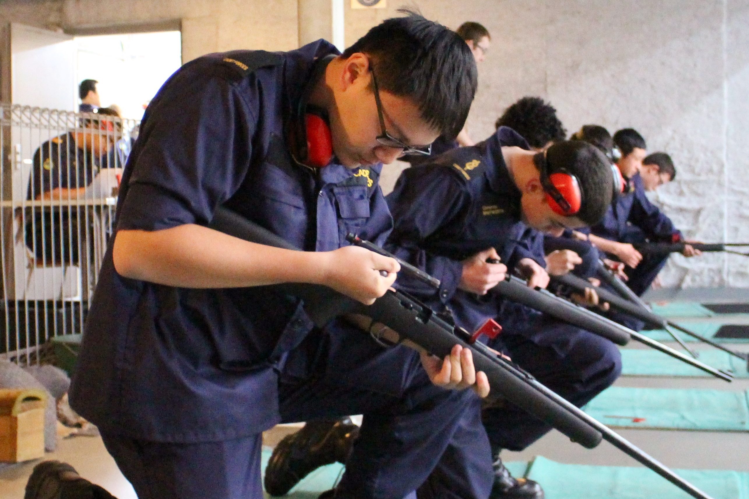 The photograph depicts an indoor target shooting drill. Individuals are arrayed in a row, donned in navy uniforms with protective ear defenders, each meticulously managing a bolt-action rifle. The nearest individual is keenly examining the bolt mechanism of their firearm. The setting is a functional indoor range with bare concrete walls and clear safety notices, underscoring the regimented atmosphere. The participants demonstrate focus and precision, indicative of a cadet training regimen.