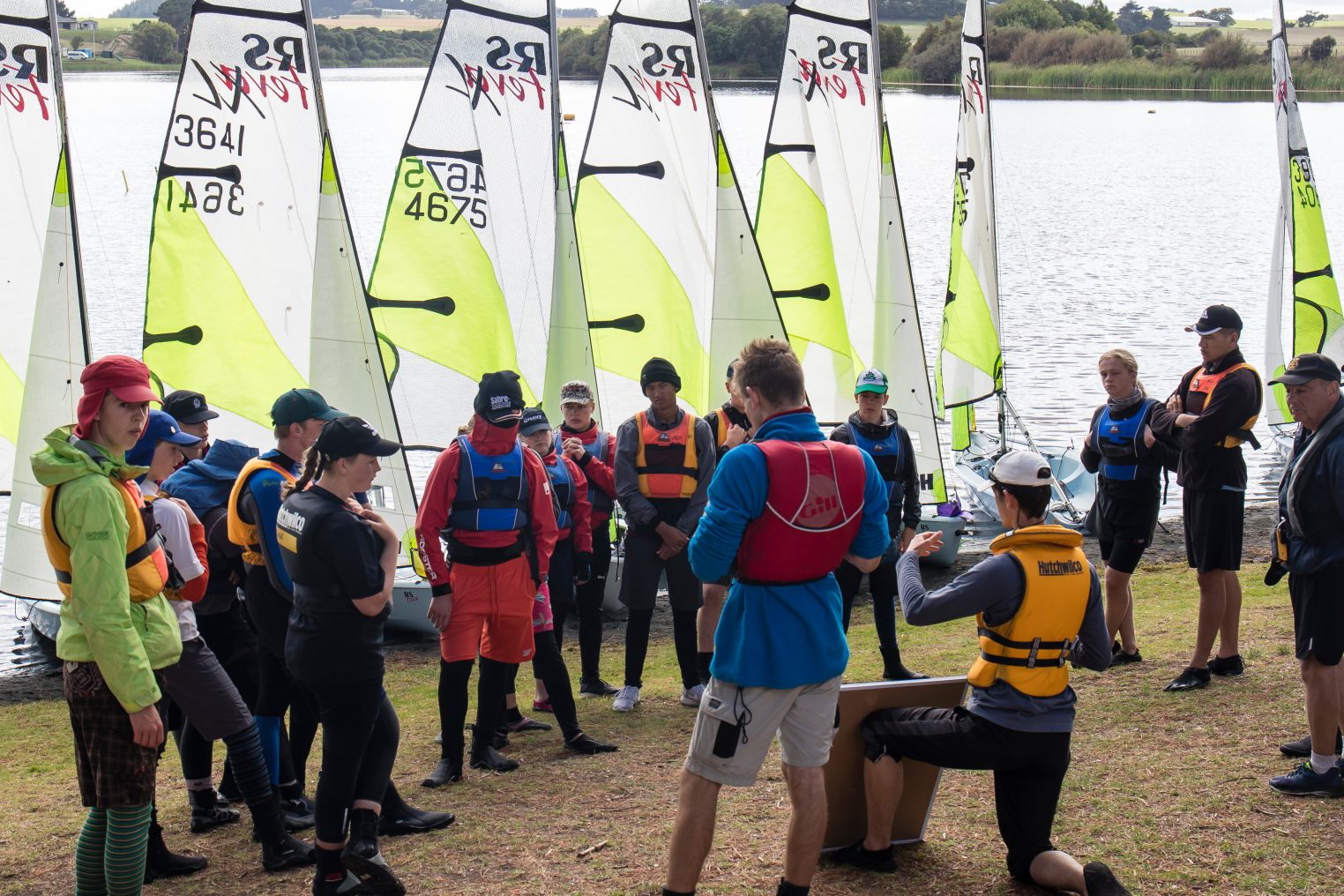 The image depicts a group of individuals at a sailing event, gathered on the grassy shore of a tranquil inland waterway. In the background, several small sailboats with white hulls and vibrant green and black sails are lined up, ready for action. The group in the foreground appears to be listening intently to a speaker, visible in a blue vest, who is addressing the team. The team members, wearing various sailing attire including life jackets in bright oranges and yellows, are attentive, some standing while others are seated or kneeling. The atmosphere suggests a briefing or instructional session before setting sail. The day is overcast, and the calm water reflects the grey sky above.