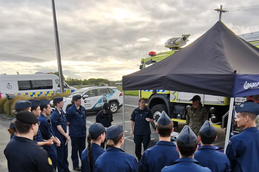 The image depicts an outdoor briefing or educational session, likely related to defense careers or emergency services. A group of individuals in navy blue uniforms with distinctive caps are attentively listening to a speaker, also in a navy uniform, who is addressing the group. The uniforms have badges that suggest they may be part of a naval cadet program or similar organization. In the background, there is a large emergency vehicle, possibly a fire truck, with the text "RNZAF FIRE" visible, indicating it belongs to the Royal New Zealand Air Force. A pop-up canopy with "NAVY" branding is also set up, under which some equipment and personnel can be seen. A police vehicle with blue and yellow checks, as well as a branded car promoting defense careers, are parked nearby, suggesting this may be a joint forces community event or a public showcase of military and emergency services. The setting appears to be in the late afternoon, as indicated by the soft lighting and the presence of shadows. The sky is overcast, with patches of blue and clouds, creating a moody atmosphere. The participants are focused and engaged, indicating the importance of the information being shared. The overall impression is one of community engagement and education about defense force careers.