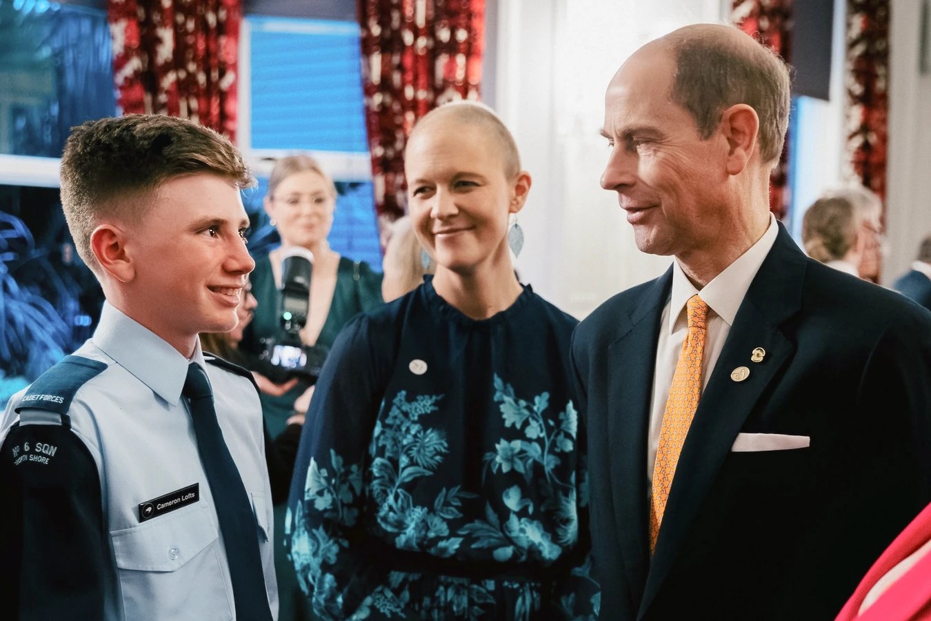 In this edited image, three individuals are engaged in conversation. In the foreground, a young man in a light blue Air Cadet uniform with a nametag reading "Cameron Lowe" is smiling, looking slightly off-camera. He appears engaged and delighted in the interaction. Standing beside him is a woman with a confident, serene expression, and wearing a dark blue dress with a light blue floral pattern. To her side, is the Duke of Edinburgh in a dark suit with a vibrant orange tie adorned with small motifs and a lapel pin is attentively listening to the young cadet. His expression is one of interest and encouragement. In the background, other indistinct figures suggest a formal event. The image evokes a sense of celebration and recognition.