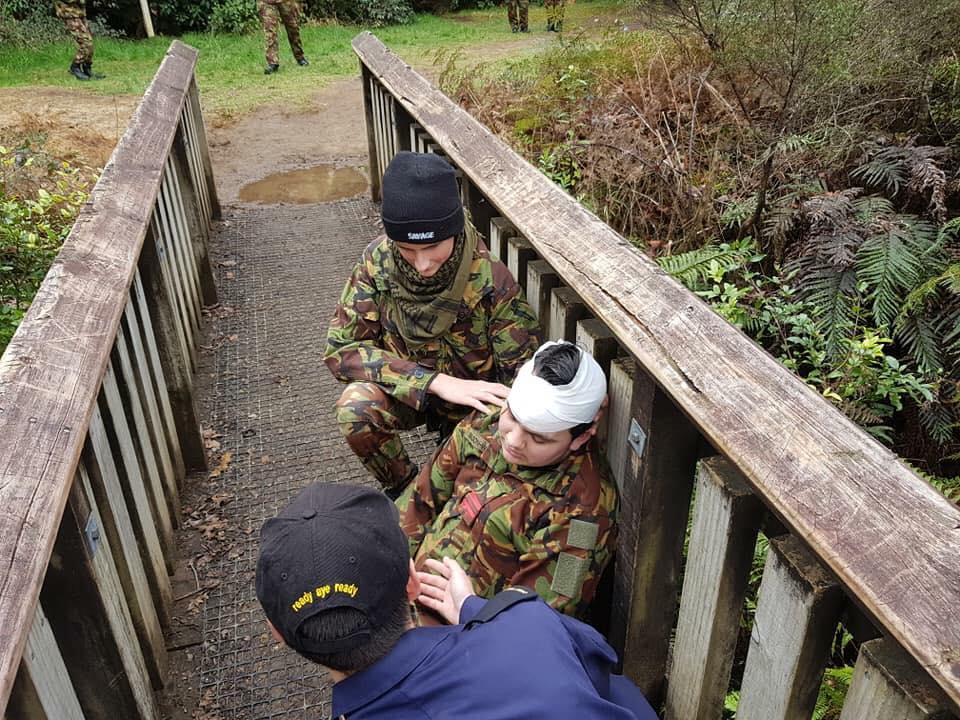 This image is set in an outdoor environment with natural foliage in the background. It captures a first-aid training scenario, where three individuals in camouflage military uniforms are attending to a fourth person seated on a wooden bridge with a handrail. One individual is carefully wrapping a white bandage around the seated person's head, suggesting treatment of a simulated head injury. Another stands by, observing the procedure, while a third looks on from a lower step, providing assistance or guidance. A black beanie with the word "SAVAGE" is visible on the head of the individual applying the bandage. Onlookers in similar military attire can be seen in the background, suggesting this is a group training exercise. The setting appears to be a forested area with walking paths, indicative of a field training environment. The focus and seriousness on the participants' faces underline the importance of the drill, emphasizing the practical skills being taught for emergency medical response in the field.