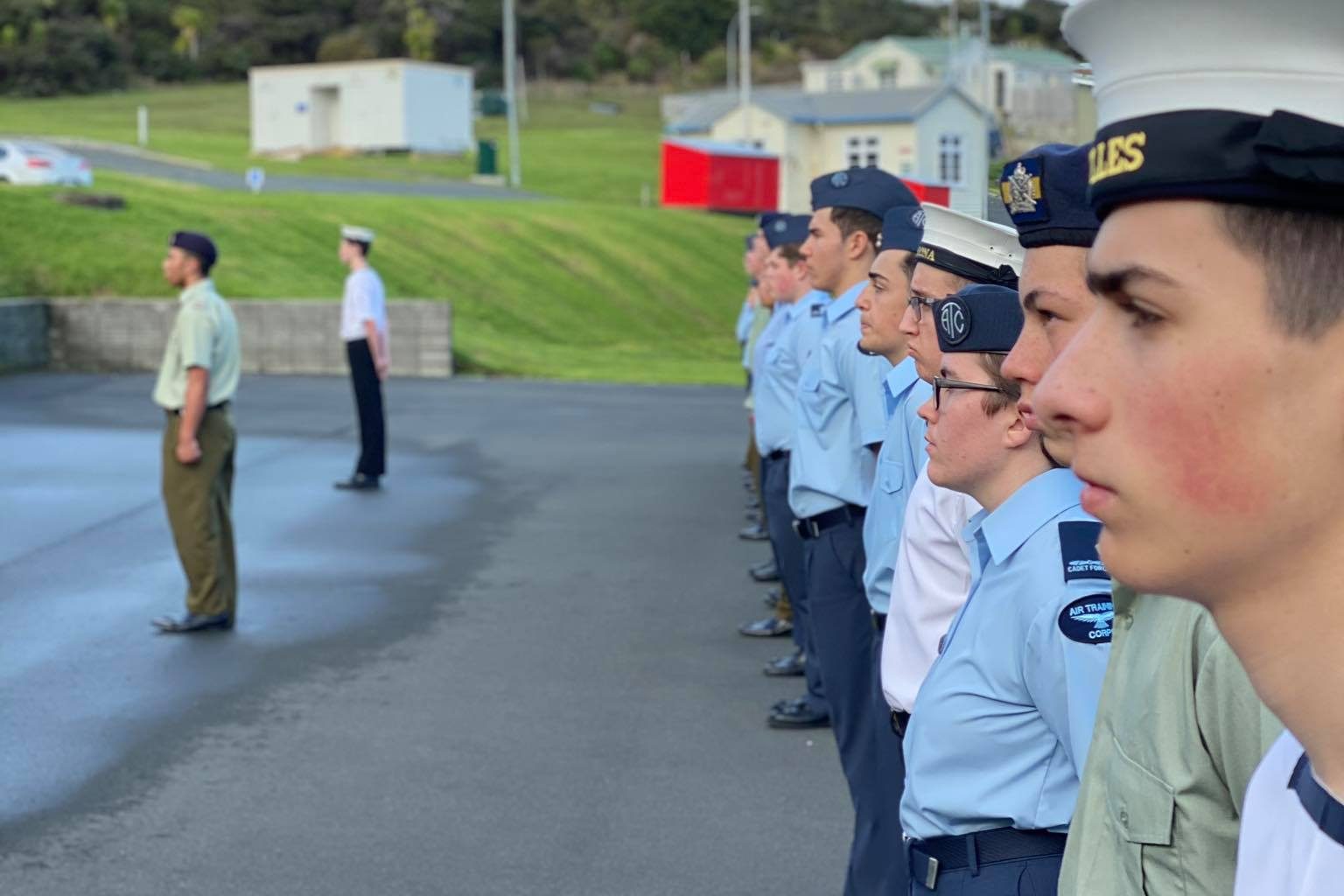 The image shows a group of young individuals in a military formation. They are dressed in light blue shirts and dark trousers, each wearing a cap with the inscription "ACHILLES". Their attire and the caps suggest they are part of a naval cadet program. They are standing at attention on a paved surface, looking off to their left. In the background, a cadet in a khaki uniform faces the group, indicating a possible drill or inspection is underway. The scene is outdoors during daytime, with green hills and a clear sky in the background, along with some buildings and cars, which gives a tranquil and disciplined atmosphere to the setting.
