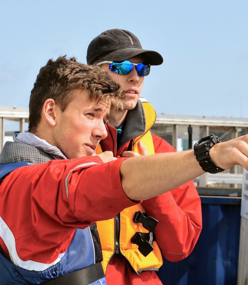 The image captures a close-up moment between two individuals engaged in an outdoor activity. The person in the foreground, wearing a red jacket with grey and white trim and a blue life vest, is gesturing into the distance, their expression intent and instructive. Behind them, a second individual, clad in a black cap and sunglasses that reflect the blue sky, is looking in the same direction with a focused gaze. Both are wearing wristwatches and appear to be involved in a navigation or orientation task. The background is out of focus, with hints of a marine setting, including the edge of a jetty and a sign that suggests proximity to water.