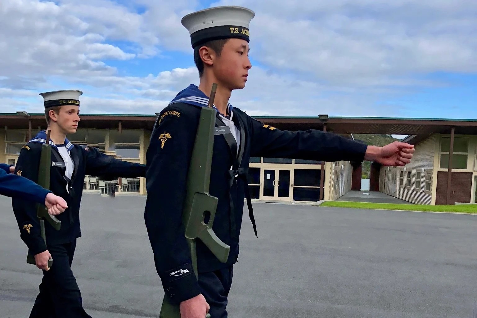 The image depicts two cadets in uniform, marching in unison on what appears to be a parade ground. The foreground is dominated by a young cadet captured mid-stride, his left arm extended in a precise, outward motion, indicative of a drill movement. He wears a dark naval cadet uniform with a white peaked cap that bears the inscription "T.S. Achilles", signifying affiliation with a particular unit or training ship. His expression is one of focused determination, with eyes fixed ahead, embodying the discipline and precision of military drill. Behind him, another cadet follows suit, mirroring the actions of the leader. The backdrop features an overcast sky and a nondescript building, placing the emphasis on the cadets and their activity.