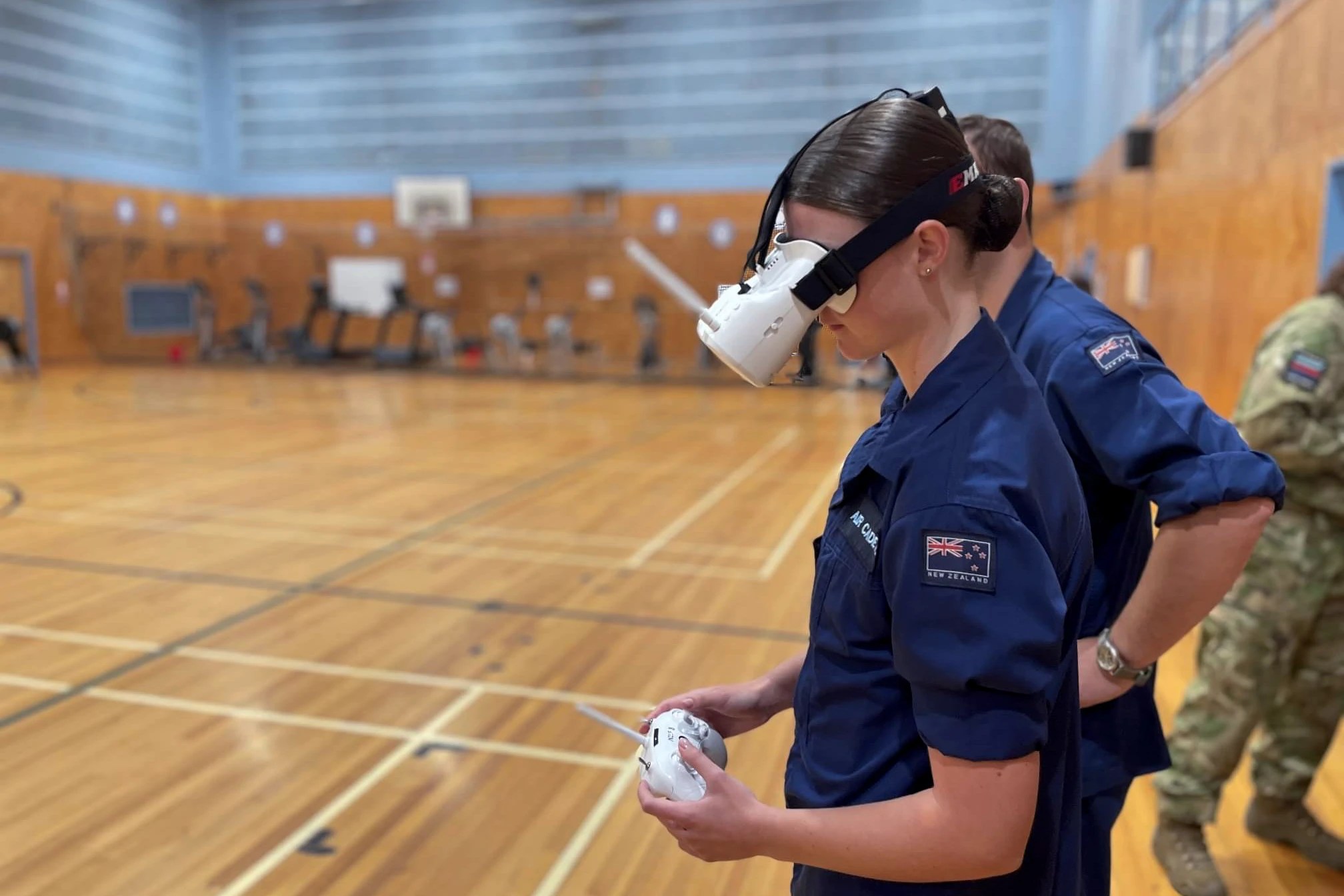 In this image, we see a young individual wearing a dark blue uniform with patches indicating "New Zealand" on the right sleeve. The person is focused on operating a white drone controller with both hands. They are wearing a white virtual reality headset that covers their eyes and is secured around their head with a black strap, which suggests they are immersed in a drone simulation experience. The backdrop is a blurred indoor basketball court, indicating an indoor setting for the NZCF Drone Operator Course. On the right, another individual in a similar uniform is partially visible, and there appears to be some activity in the background, although it is out of focus. The environment suggests a practical, hands-on learning scenario.