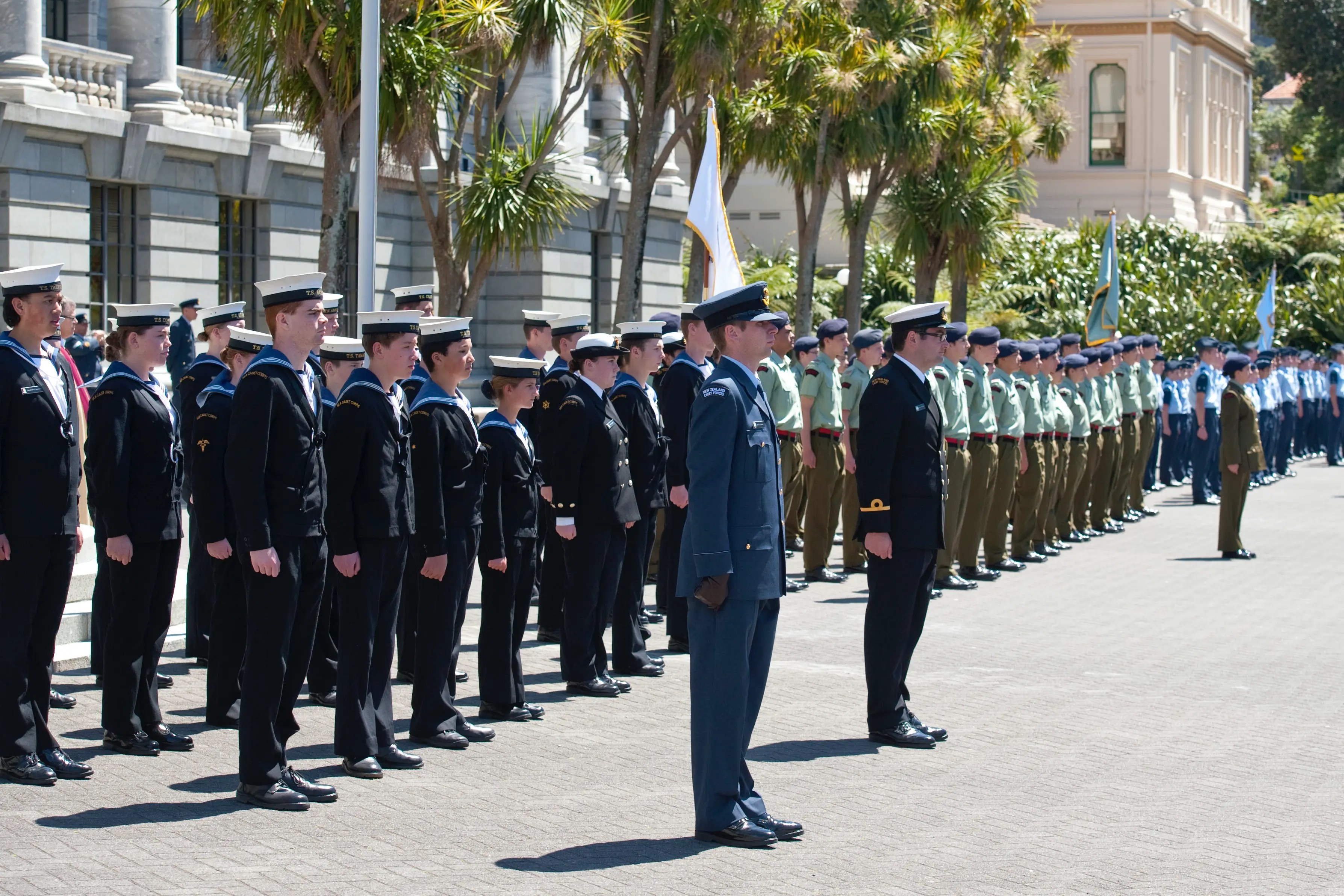 The image captures a formal military parade under clear blue skies. It features uniformed personnel from various branches lined up in formation. The first two rows are dressed in navy blue uniforms with white peaked caps, likely representing naval forces. Following them are individuals in light blue uniforms with darker caps, indicative of air force personnel. The subsequent rows are filled with individuals in olive green uniforms, typical of army attire. Each member stands erect and attentive. Behind them, the architecture suggests a formal, possibly governmental, setting with palm trees dotting the landscape, adding a serene tropical feel to the disciplined scene. The atmosphere is one of solemnity and pride, reflective of the importance and dignity of the occasion.