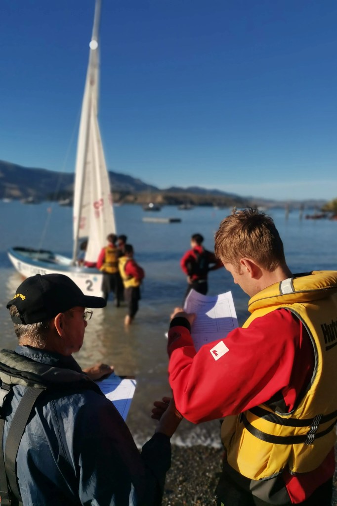 The image captures a serene, sunny day at a lakeside with clear blue skies. In the foreground, two individuals are engaged in a discussion. One, wearing a dark cap and a navy jacket with a backpack, is holding papers and discussing their content with the other person, who is wearing a bright red life jacket with yellow elements, suggesting they are a participant in a water-based activity, possibly sailing. The focus is on these two individuals, with the background slightly blurred. Behind them, several people, potentially other crew members or participants, are in the shallow waters near the shore, with a white sailboat visible, numbered '2'. The calm waters of the lake gently lap at the shore, and other boats can be seen in the distance, along with a mountain range that creates a majestic backdrop. The interaction between the two individuals in the foreground suggests a briefing or a review of the sailing activity, possibly discussing safety or instructions. The attire of the person in the life jacket indicates they are ready for an aquatic activity. The overall atmosphere is one of preparation and anticipation for sailing on this picturesque day.
