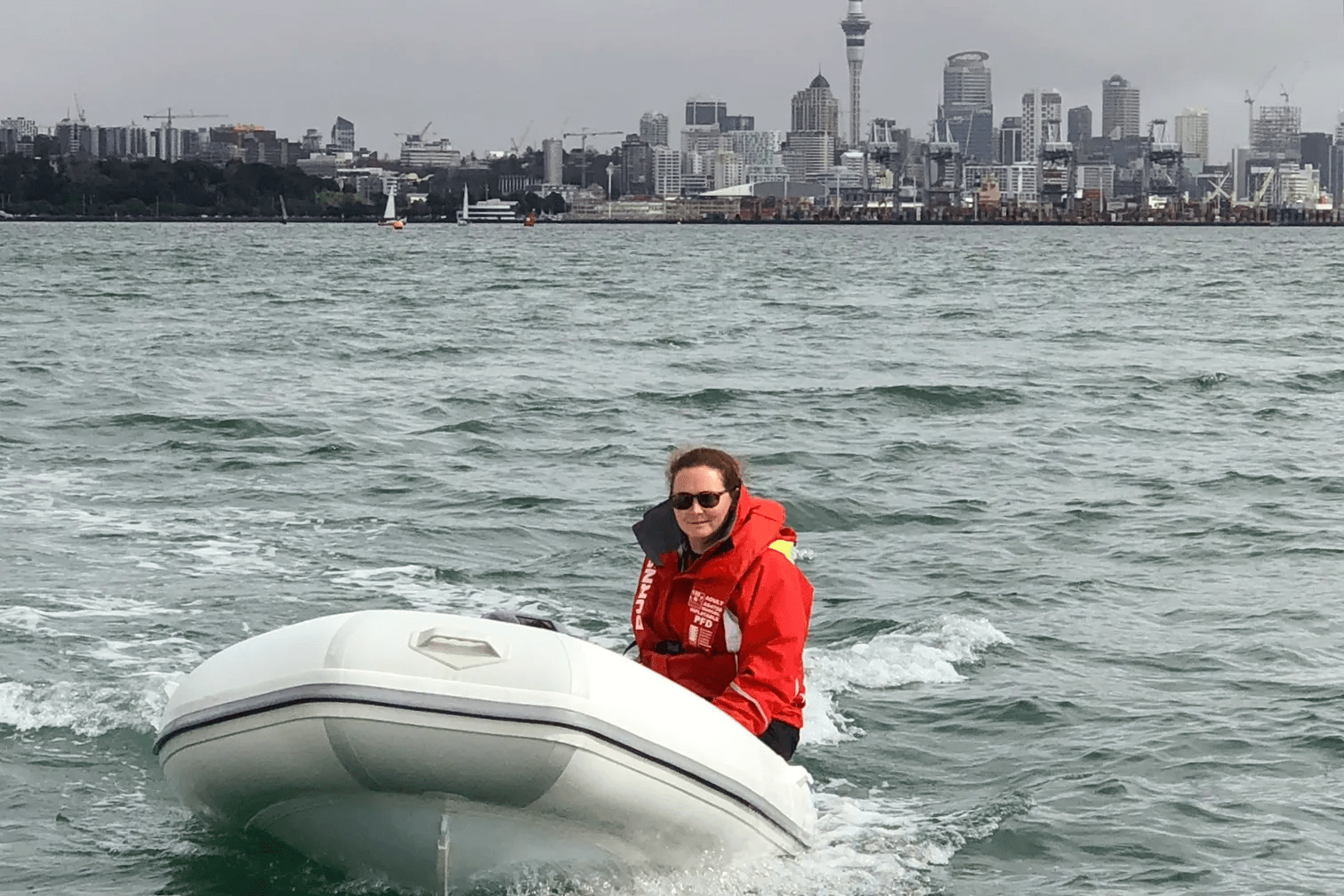 This image shows a person driving a rigid-hulled inflatable boat (RHIB) on choppy water with a city skyline in the background. The individual is dressed in maritime safety gear, including a bright red life jacket and sunglasses, suggesting preparedness for water activities. The RHIB appears to be moving at speed, with water spray around the sides. Behind, the skyline features a variety of buildings and a tall tower, indicative of a large city. The overcast sky suggests it might be a cool day. The person is seated towards the rear of the boat, with their hands presumably on the controls, which are not visible in the photo. The focus on the individual and the RHIB, along with the expansive view of the city, gives the sense of an active moment in a busy, urban waterfront setting.