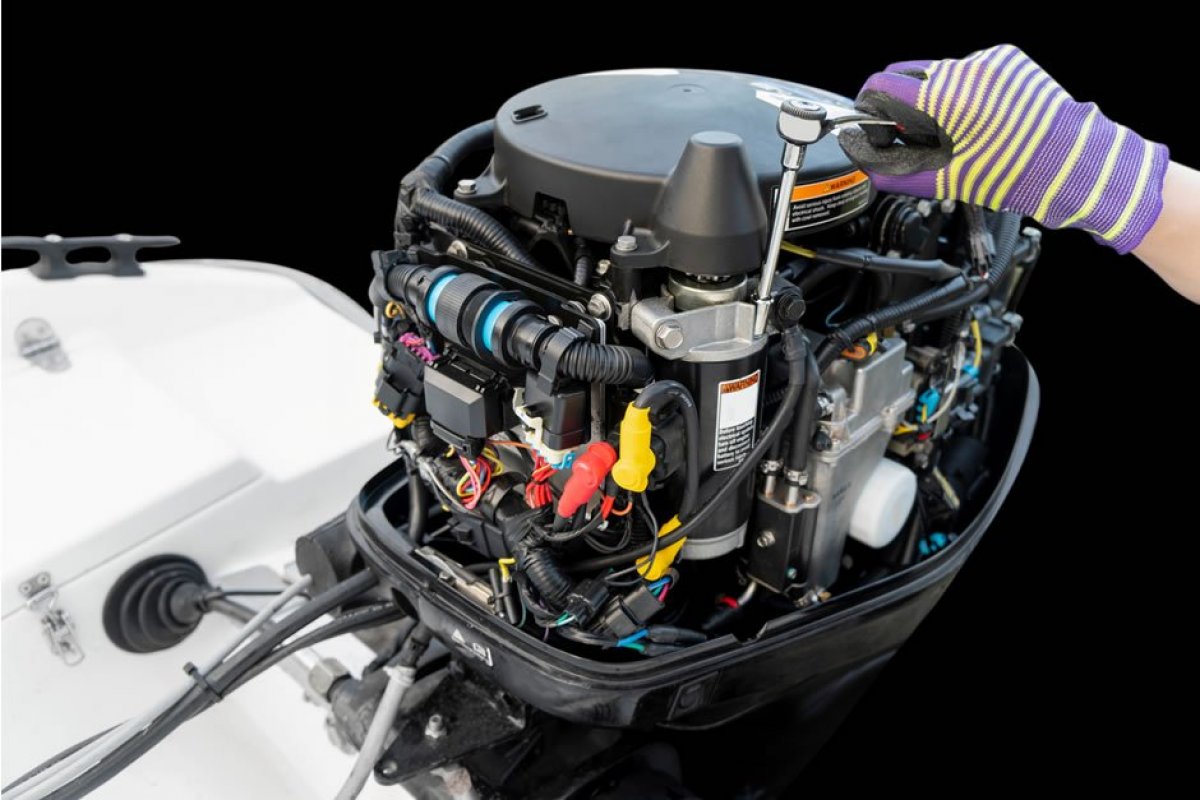 This image features a close-up of a modern outboard boat engine, with someone's hand in the foreground wearing a striped purple and grey glove, working on the engine. The engine is detailed and complex, with various components such as electrical wiring in multiple colors, black ribbed hoses, metallic cylinders, and parts with labels and warning stickers visible. The engine cover is a dark grey color and the engine itself contains parts in black, silver, and with color-coded elements like red and yellow caps, suggesting different functions or fluid types. The engine is mounted on the back of a white boat, and the background is a blurred black void, focusing the viewer's attention on the engine and the maintenance work being performed. The overall image portrays a maintenance or inspection process, indicative of regular upkeep and attention to the intricate systems that enable the motor's operation. The clarity and depth of the components suggest a well-maintained piece of equipment, essential for the reliability and safety of marine transport.