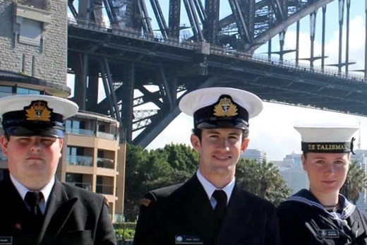 The image features three individuals in naval uniforms, with the iconic Sydney Harbour Bridge in the background, suggesting the location is Sydney, Australia. The two outer individuals are wearing peaked caps with the badge of the New Zealand Cadet Forces, while the person in the middle has a cap tally with "TS TALISMAN" inscribed, indicating they are part of a sea cadet corps, likely from New Zealand given the badge design. All three are wearing dark navy double-breasted jackets with brass buttons, white peaked caps, and black neckties. The central figure stands out with additional decorations: two thin gold stripes on their sleeves indicating a higher rank, possibly that of a junior officer. Their posture is attentive and formal, reflecting the disciplined nature of cadet or naval service. The image captures a moment of pride or ceremonial significance, possibly during an international cadet exchange or a public event. The setting is bright and clear, and the presence of such a well-known landmark provides a strong sense of place and occasion.