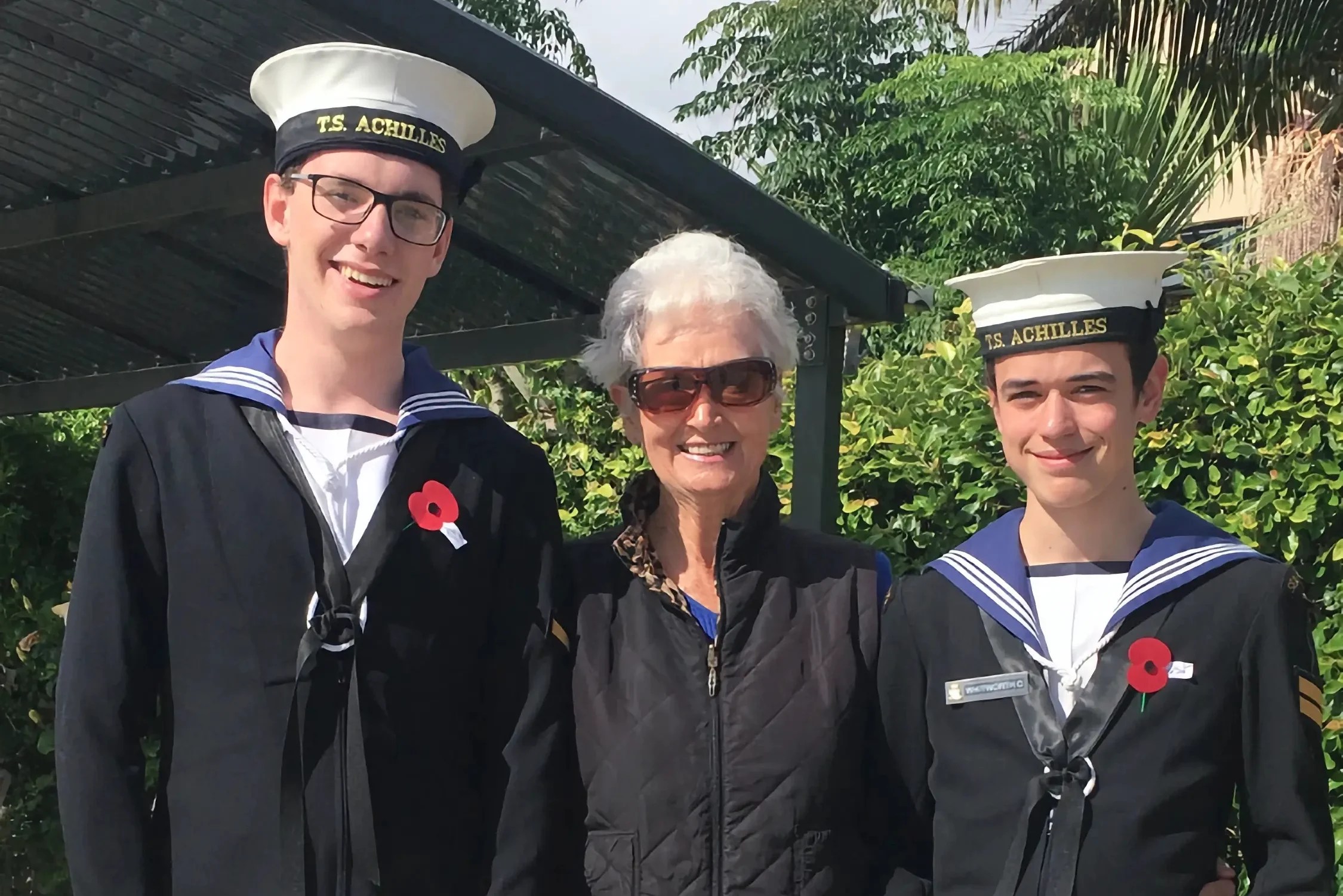 The image features two cadets and an older woman standing outside on a sunny day. The cadets are dressed in naval cadet uniforms, with white hats labeled "T.S. Achilles" and dark jackets with white belts. Both have red poppies pinned on their jackets, indicating the photo was likely taken around Anzac Day or Remembrance Day. The cadet on the left holds a collection box labeled "RSA," which stands for the Returned and Services' Association, an organization supporting military veterans in New Zealand. The cadet on the right holds a donation bucket. The woman in the center, likely a supporter or perhaps a veteran herself, wears a black puffer vest, black trousers, and sunglasses, with a poppy on her vest. They are all smiling, suggesting a positive atmosphere during a commemorative event. The setting includes a covered walkway and a backdrop of trees and a residential area. The image captures a moment of intergenerational connection and the continuation of traditions honoring military service.