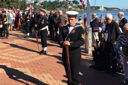 This image shows a sunny day at a commemorative event, most likely ANZAC Day, at what appears to be a waterfront location. In the foreground, a person in naval uniform stands at attention, holding a ceremonial rifle vertically in front of them with both hands. They are wearing a white peaked cap, a navy jacket with medals affixed, white gloves, and dark trousers with a stripe down the side. Behind them, two flags are at half-mast on a flagpole, the New Zealand flag and another flag with Union Jack at the corner and a Southern Cross, indicating an Australian connection. A crowd of onlookers, some of whom are wearing poppies, watches the ceremony with respect and attention. The location, suggested by the presence of water and boats in the background, could be a seaside memorial in New Zealand. The atmosphere is solemn and respectful, befitting a day of remembrance.