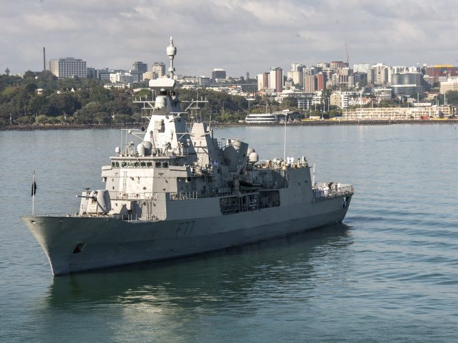 The image captures a large naval frigate with the hull number F77 prominently displayed on its bow. The ship is gray with various levels of decks, antennas, and a large superstructure. Crew members can be seen on the decks, suggesting activity and readiness. The vessel is afloat in calm waters with a city skyline in the background, featuring buildings of different heights, trees, and a clear sky above. The setting suggests a peaceful port city, and the ship is likely either departing or arriving at the harbour. The overall impression is one of a modern maritime force within an urban coastal setting.