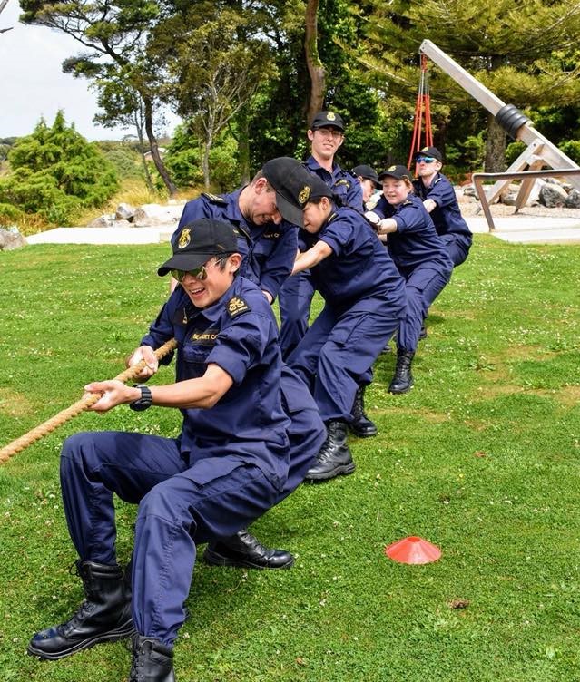 This image captures a moment of high spirits and teamwork during an outdoor tug-of-war contest. A group of people in dark navy uniforms, identified by patches as members of a cadet corps, are in the midst of a pull. They are leaning back, digging their heels into the grass to gain traction. The focus is on the front person who is grinning, holding the rope tightly, and seems to be leading the effort. They are wearing utility caps, and one of them is wearing glasses. The scene is set on a grassy field with a cannon and trees in the background, suggesting a park or recreational area. The weather appears pleasant, with a mix of sun and clouds.